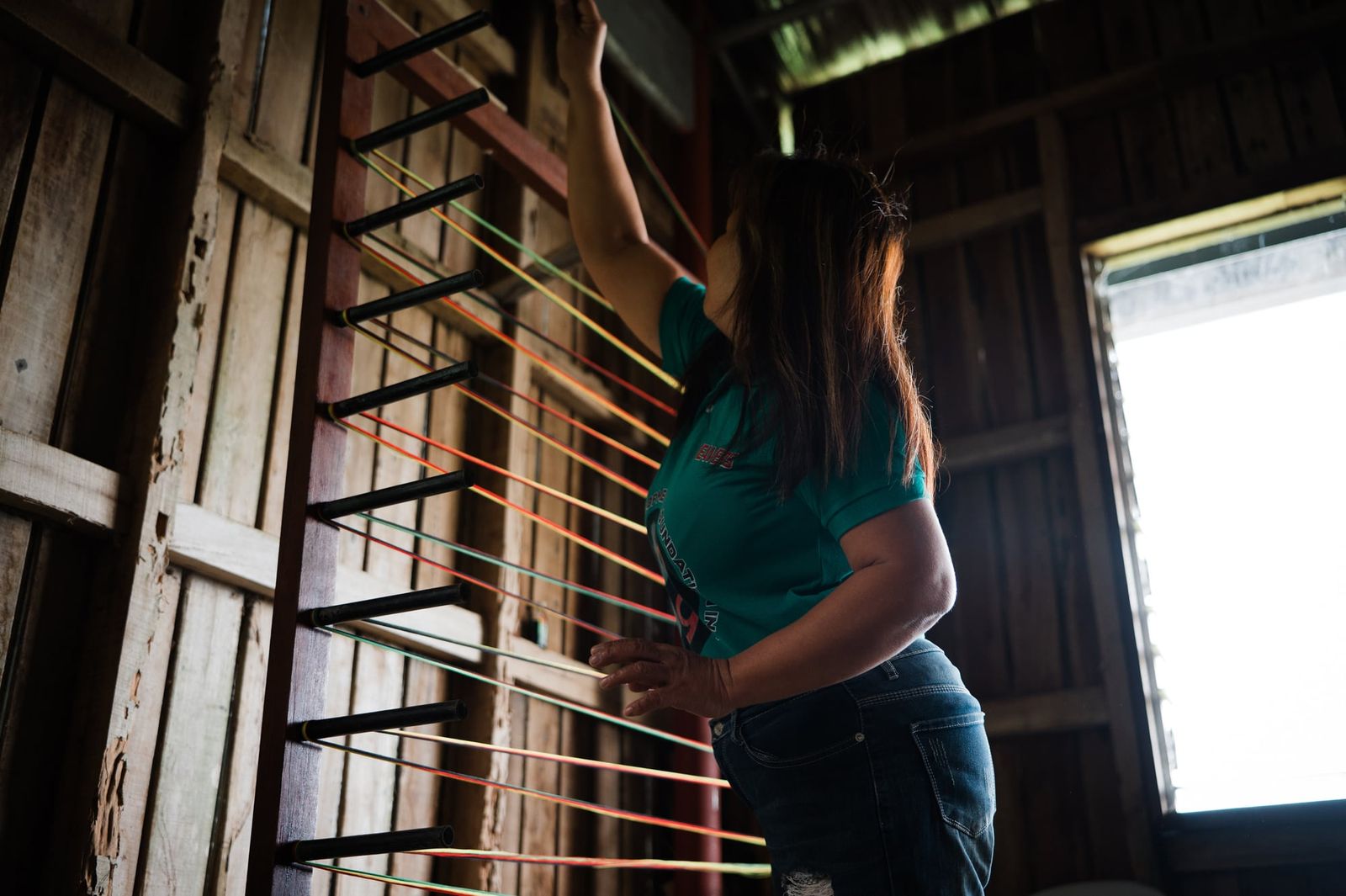 A weaver reaching up to wind thread on the warping frame by the window
