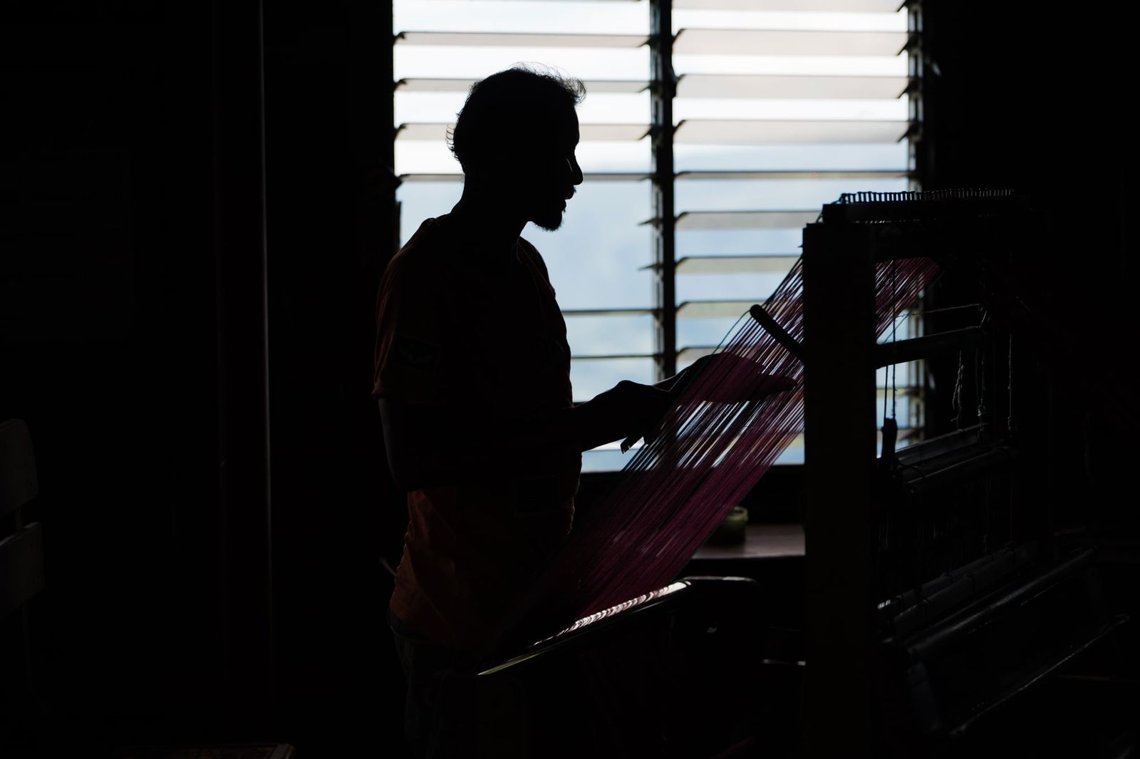 A weaver in silhouette against the window, working at the loom
